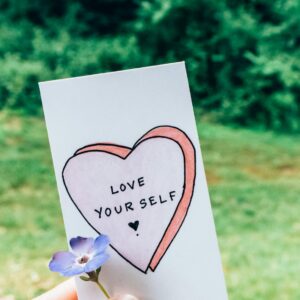 A hand holds a card with 'Love Yourself' next to a small flower outdoors.